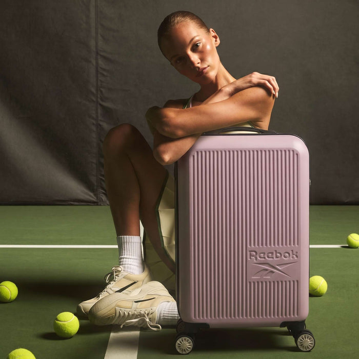 A woman in tennis attire sits on a court, arms resting on a pink Reebok Action Duo 2 Piece Luggage Set with spinner wheels. Tennis balls and a racket are scattered nearby on the green surface.