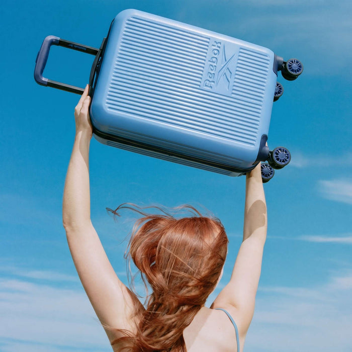 A person with long, reddish hair lifts a blue Reebok Action Duo 2 Piece Luggage Set overhead against a bright blue sky with wispy clouds.