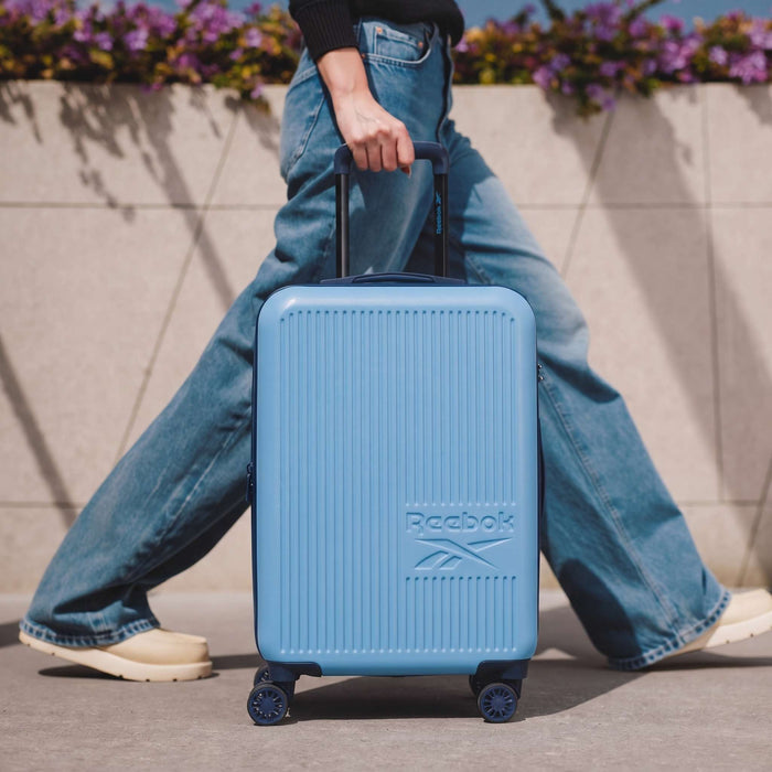 Wearing wide-leg blue jeans and cream shoes, a person pulls a light blue Reebok Action Duo 2 Piece Luggage Set outdoors near a wall with flowers.