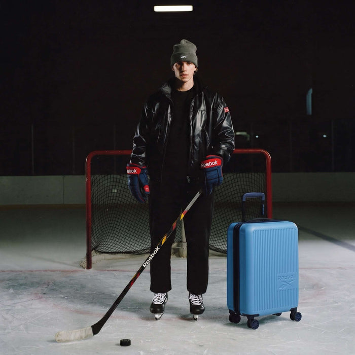 A person in a leather jacket, beanie, hockey gloves, and skates stands on an ice rink with a hockey stick, puck, and the Reebok Action Duo 2 Piece Luggage Set featuring spinner wheels.