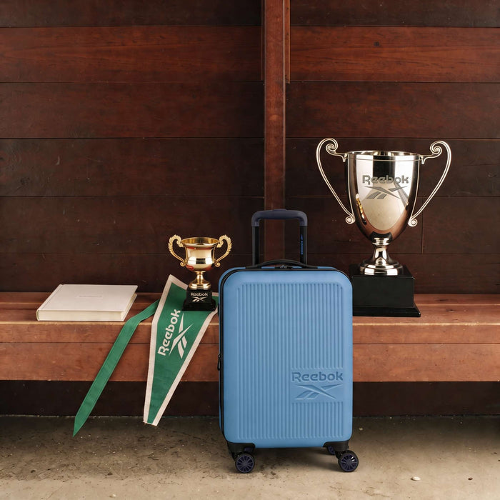 A blue Reebok Action Duo 2 Piece Luggage Set with spinner wheels rests on a wooden bench next to two trophies, a green Reebok pennant, and a closed white box, all set against a wooden wall.
