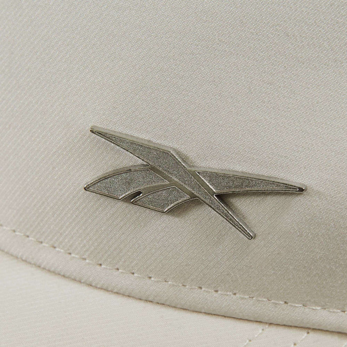 Close-up of the Reebok Badge Cap shows its beige fabric surface with a metallic angular Reebok logo and visible stitching along the edge.