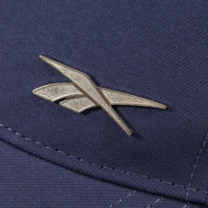 A close-up of a metallic Reebok logo pin on the Badge Cap, featuring dark blue fabric with detailed stitching and a shiny logo.