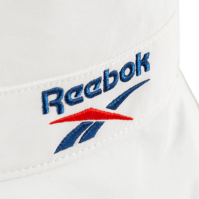 Close-up of a white Reebok Bucket Hat, made from recycled nylon, showcasing the blue and red embroidered Reebok logo above a stylized vector—a striking streetwear piece.