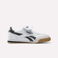 The Campio XT Kilty Shoes by Reebok feature a white and black leather upper, gum sole, tennis-inspired style, black Reebok logo, and Union Jack flag detail on the side. Shown against a plain white background.