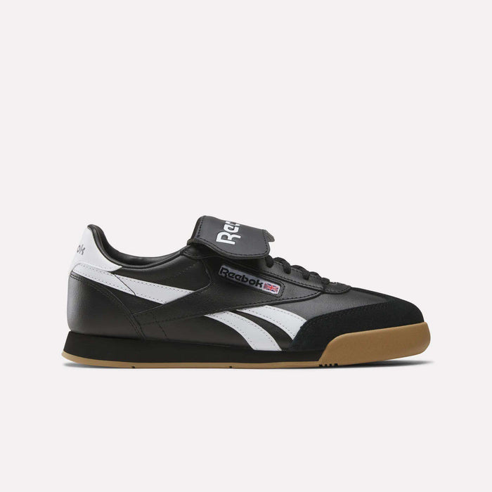 The Reebok Campio XT Kilty Shoes are black sneakers with white side stripes, a gum sole, Velcro strap, tennis-inspired style, classic leather construction, the Reebok logo, and a British flag detail on the side.