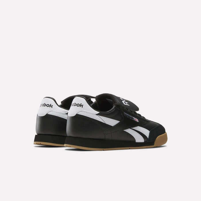 A pair of Reebok Campio XT Kilty Shoes in black with white stripes, white heel accents, gum soles, and velcro straps are displayed on a light background, angled to showcase the back and side tennis-inspired design.