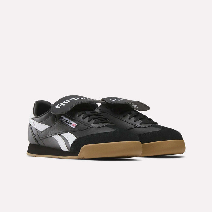 The Reebok Campio XT Kilty Shoes feature black uppers, white side stripes, gum soles, black laces, and wide black Velcro straps for a tennis-inspired look, shown on a plain white background.