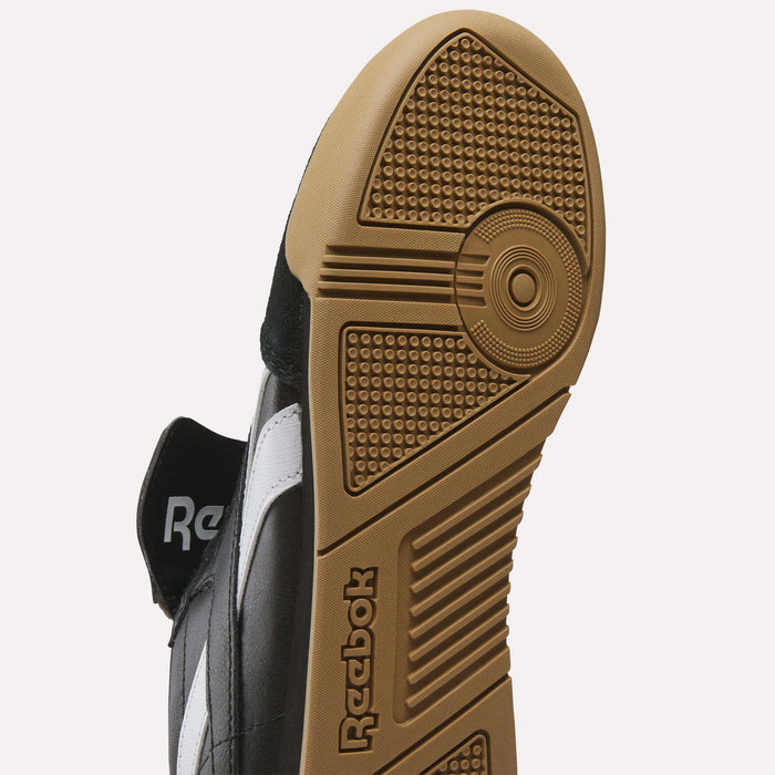 Close-up of the Reebok Campio XT Kilty Shoes' sole, featuring a textured brown rubber outsole with circular and rectangular patterns, the Reebok logo, and part of the black and white upper for a tennis-inspired look.