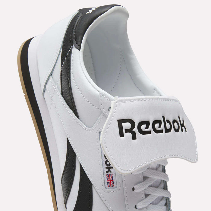 A close-up of white leather Reebok Campio XT Kilty Shoes with black accents, showcasing the Reebok logo on the tongue and a small Union Jack flag on the side. The tennis-inspired style stands out with the tongue pulled forward, revealing the inside.