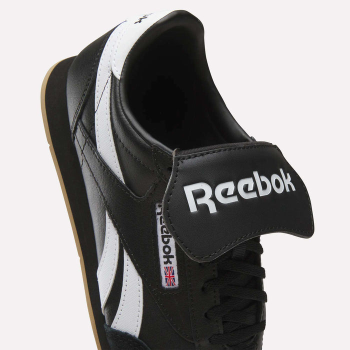 A close-up of the Reebok Campio XT Kilty Shoes in black leather with white stripes, featuring tennis-inspired style, a bold Reebok logo on a large tongue flap, and a small UK flag tag on the side.