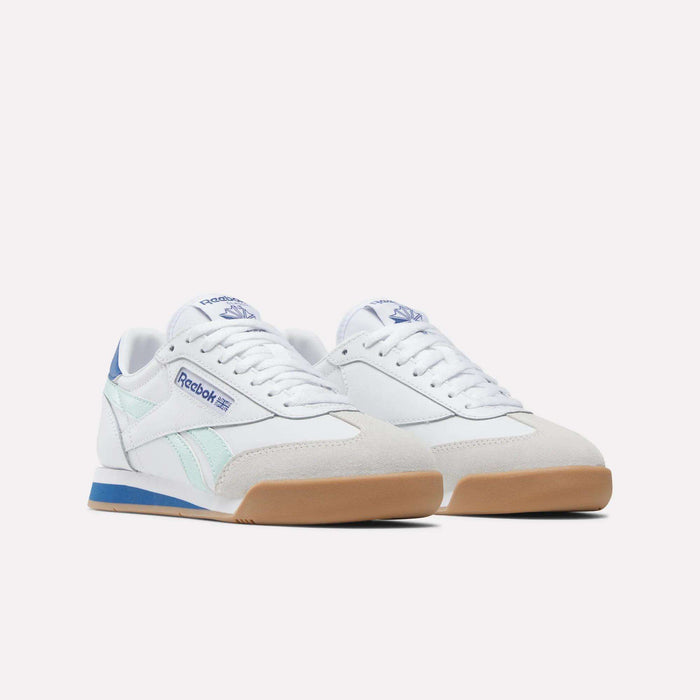 A pair of Reebok Campio XT Shoes - Big Kids featuring white leather uppers, light blue and teal retro soccer-inspired side stripes, blue branding, white laces, and gum rubber soles are displayed on a plain light background.