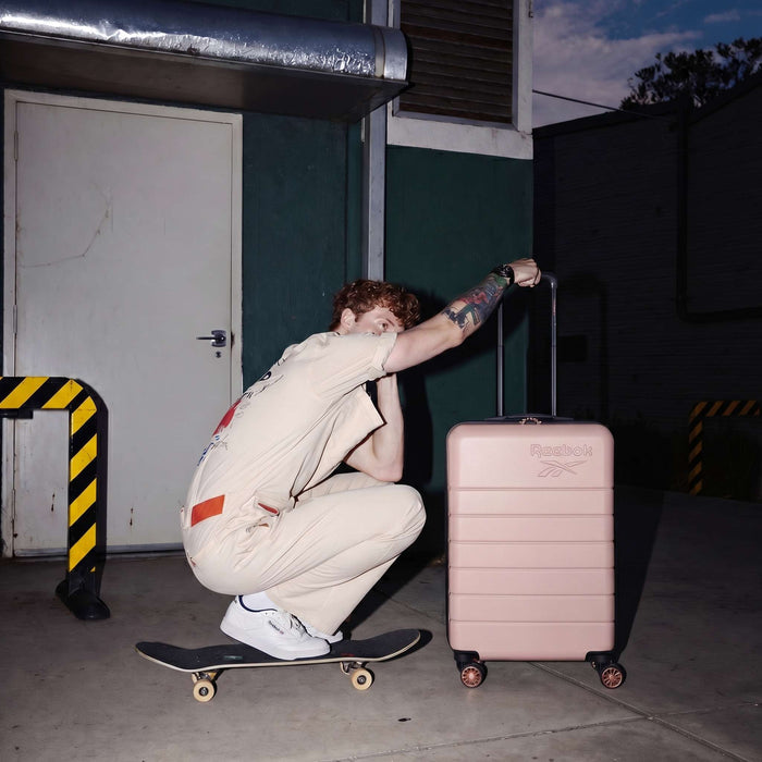 A tattooed person in a light-colored jumpsuit crouches on a skateboard at dusk, holding the handle of the pink Reebok Captain 2 Piece Luggage Set with spinner wheels. They wear white sneakers and are outside a building.
