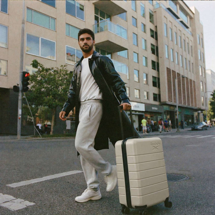 Wearing a long black coat, white sneakers, and gray sweatpants, a man crosses a city street with the Reebok Captain 2 Piece Luggage Set. Modern buildings and pedestrians fill the background as he pulls his spinner-wheeled suitcase.
