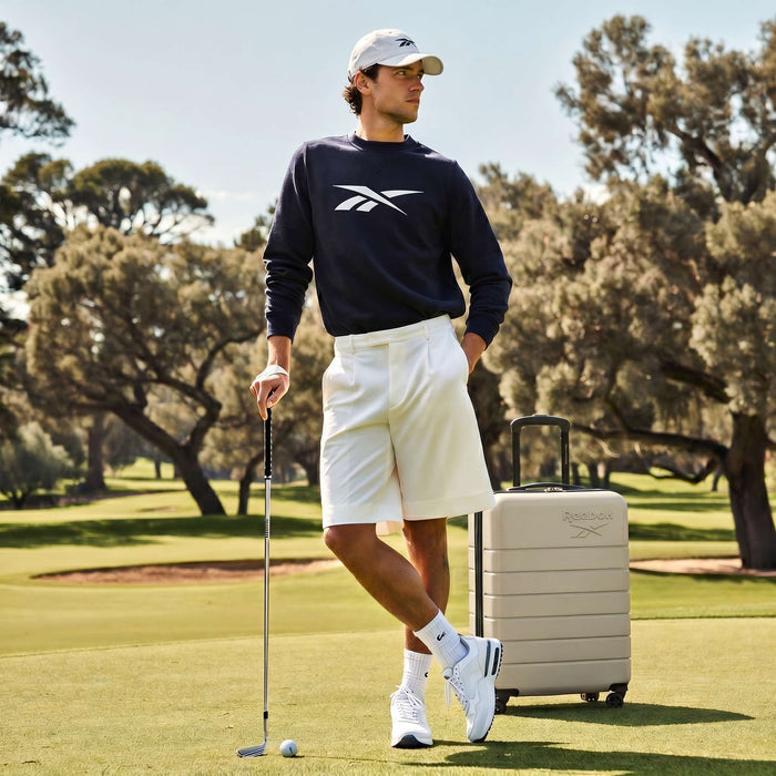 A man in a dark sweatshirt, white shorts, sneakers, and cap stands on a golf course holding a club beside Reebok’s Captain 2 Piece Luggage Set with spinner wheels, surrounded by trees and green grass.