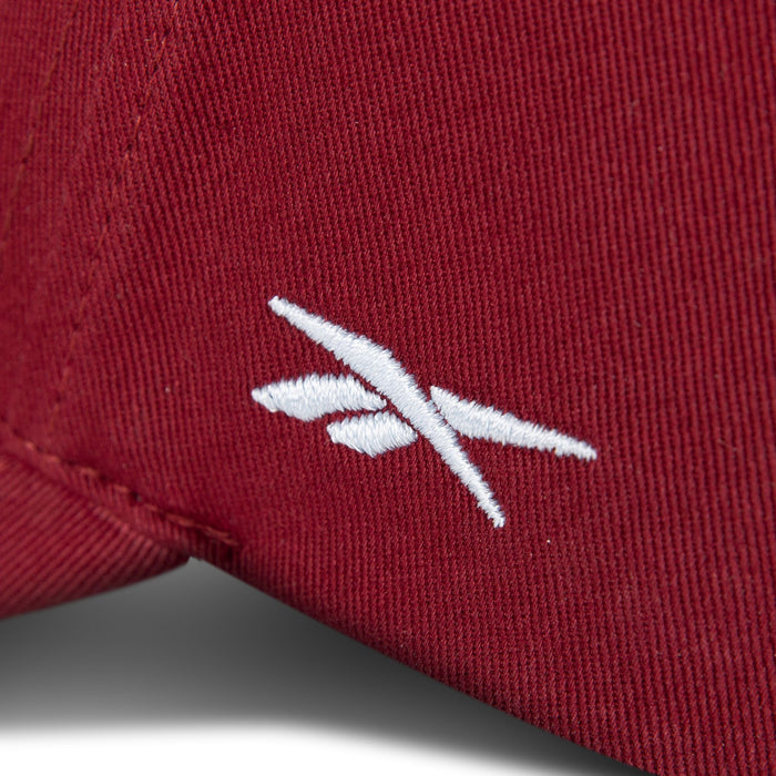 Close-up of the Reebok Chroma Cap, an adjustable cotton twill snapback. The maroon cap features detailed stitching and a small white embroidered abstract logo with two intersecting lines on the textured fabric.