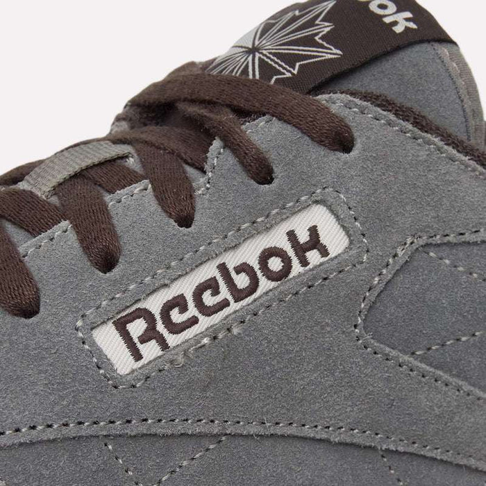 Close-up of the Reebok Classic Leather Premium LTD Shoes in gray suede, featuring brown laces, intricate stitching, and the Reebok logo displayed on a fabric label. A portion of a star-shaped design appears on the tongue, emphasizing the iconic silhouette of these retro-inspired sneakers.