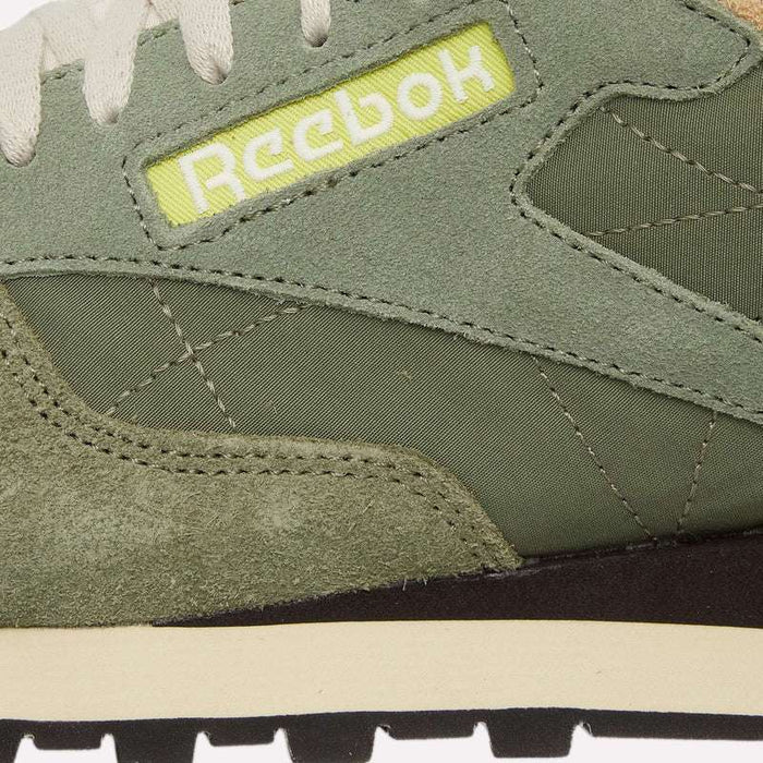 Close-up of a green Reebok Classic Leather Premium LTD Shoe, highlighting the retro charm of its classic silhouette with detailed stitching, a mix of suede and canvas textures, white laces, and the yellow Reebok logo on the side. The black and white outsole is also partially visible at the bottom.