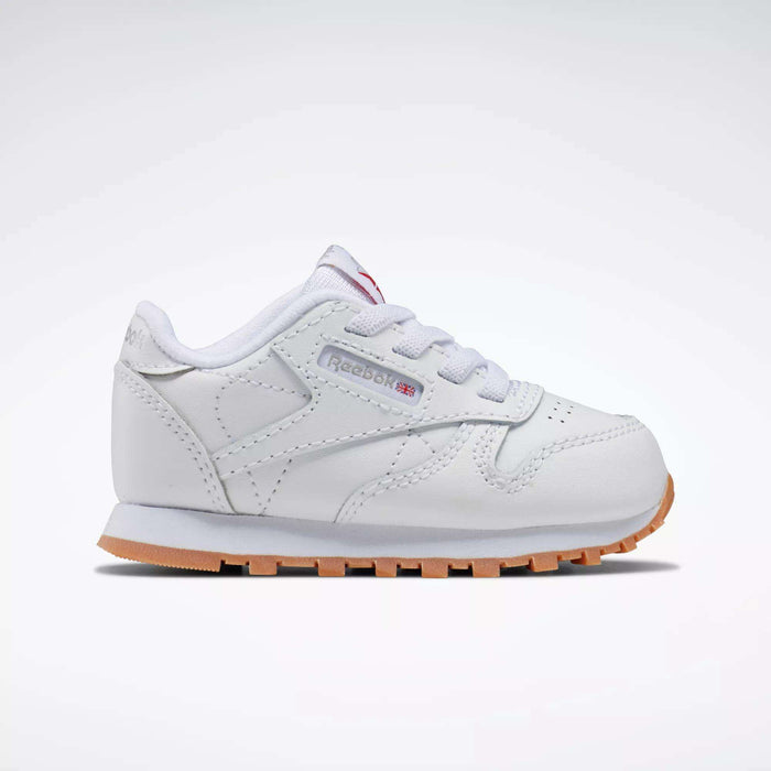The Reebok Kids' Classic Leather Shoes for babies and toddlers are white with a light brown sole, white laces, and a side tag featuring the Reebok logo, shown from the side on a plain white background.