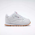 The Reebok Kids' Classic Leather Shoes for babies and toddlers are white with a light brown sole, white laces, and a side tag featuring the Reebok logo, shown from the side on a plain white background.