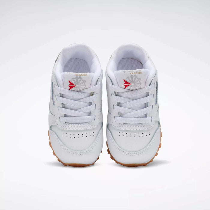 The Reebok Kids' Classic Leather Shoes - Baby & Toddler feature white leather, gum-colored soles, white laces, and a red and gray starburst logo on the tongue—perfect for active toddlers.