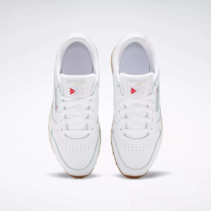 A pair of white Reebok Classic Leather Shoes for big kids, with gum soles and stitched side stripes, viewed from above. Red accents on the tongue stand out as the shoes are positioned side by side on a light gray background.