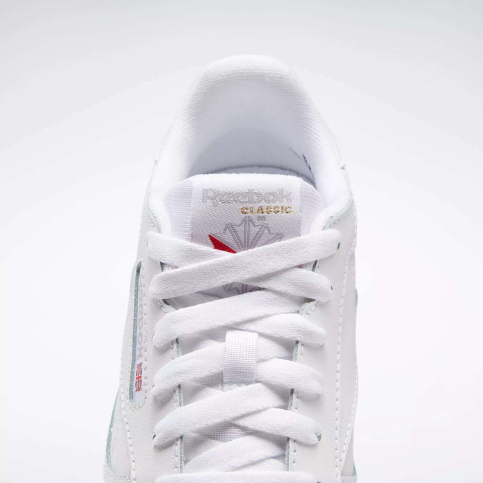 A close-up of the Reebok Classic Leather Shoes - Big Kids highlights their timeless white design, showing the laces, tongue, and a gold Reebok logo with a red emblem above it.