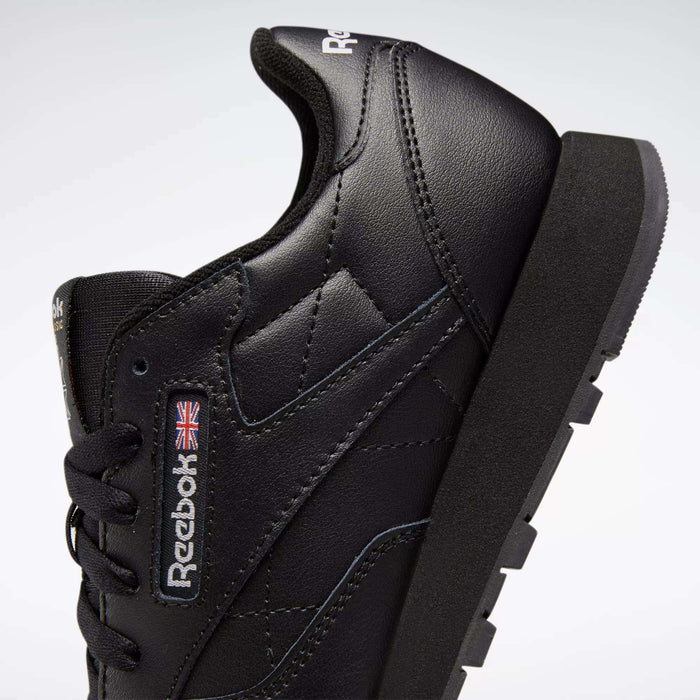 A close-up side view of the Reebok Classic Leather Shoes - Big Kids in black, showcasing the iconic design with stitched leather details, Union Jack flag, and Reebok logo—ideal for stylish boys' footwear.