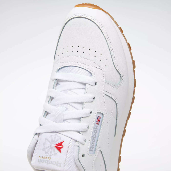 A close-up of the Reebok Classic Leather Shoes - Big Kids in white, featuring a gum sole, logo, red and gray accents, stitched side-stripe, perforated toe box, and white laces.
