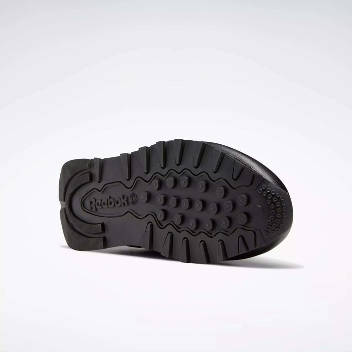 The sole of these Classic Leather Shoes - Big Kids by Reebok, shown in black from the bottom, has textured grip patterns and an embossed Reebok logo near the heel, emphasizing a timeless design.