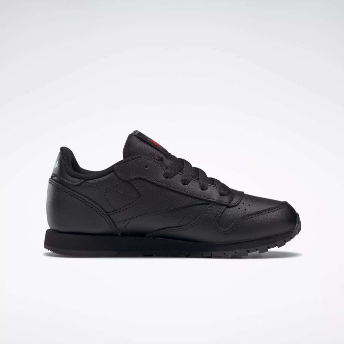 The Reebok Kids' Classic Leather Shoes - Preschool feature a black low-cut sneaker design with flat black laces, a textured rubber sole, and lightweight cushioning for comfort, shown against a plain white background.