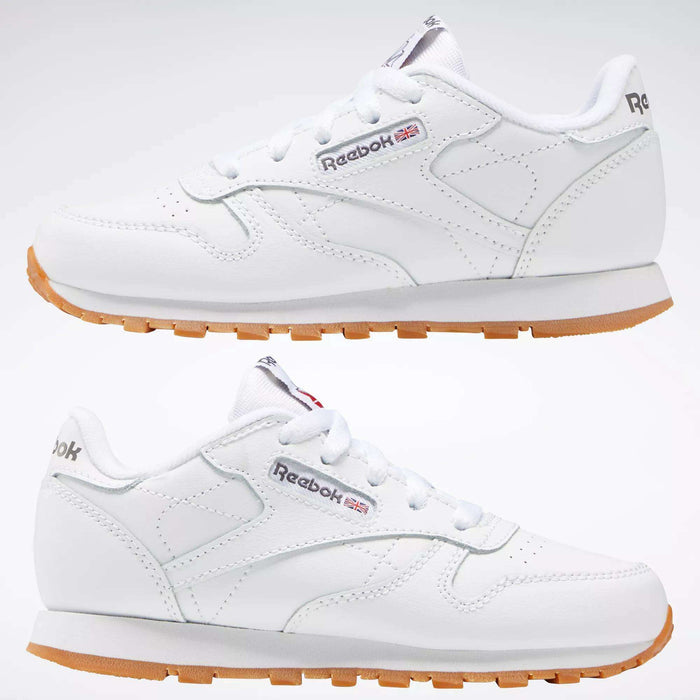 Two Kids' Classic Leather Shoes by Reebok in white are displayed side by side, showing the outer and inner sides. They feature a leather upper, Reebok branding, subtle stitching, and lightweight cushioning for comfort.