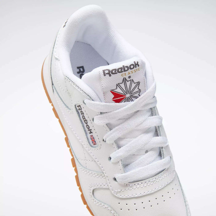 A close-up of Kids' Classic Leather Shoes - Preschool by Reebok, featuring a white leather upper, white laces, gum sole, Union Jack Reebok logo on the side, and a branded tongue with starburst design.