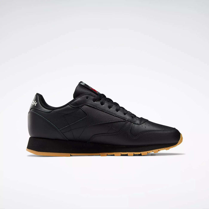 Side view of Reebok Classic Leather Shoes in black with a gum sole, black laces, minimal stitching, and low-cut design. These retro-inspired shoes have a durable leather upper and are shown on a plain light gray background.