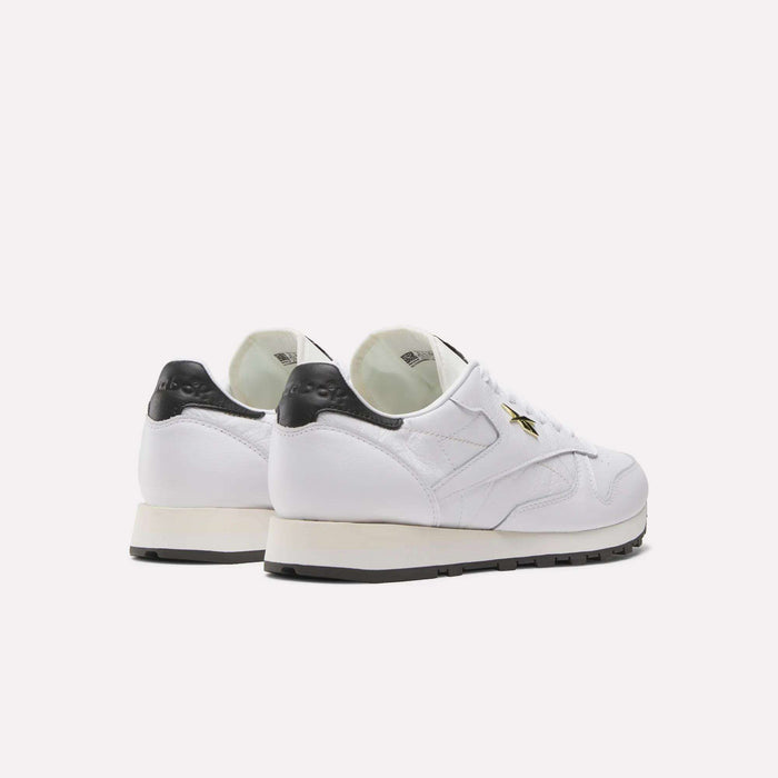 The Reebok Classic Leather Shoes feature a white leather upper, black heel accents and soles, plus a gold star detail on the side, reflecting retro running style. Displayed on a plain white background.