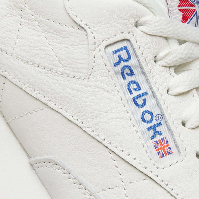Close-up of Reebok Classic Leather Shoes shows the blue Reebok logo and a small Union Jack on the durable white leather side panel, with white laces highlighting their retro style.