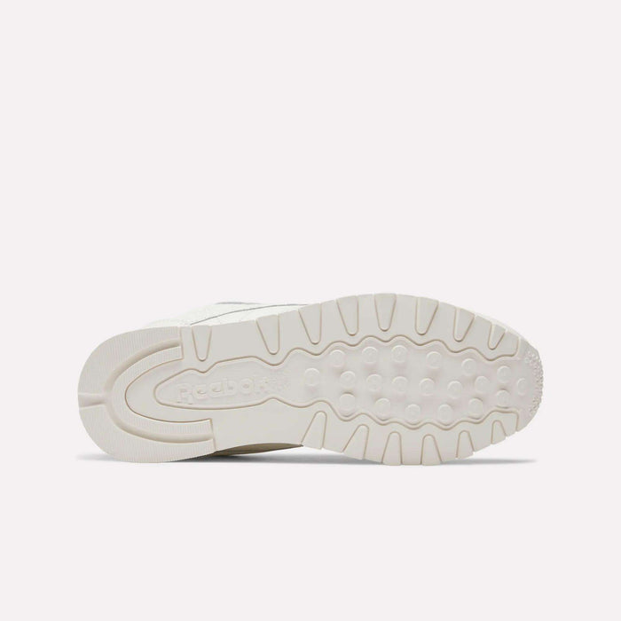 The image shows the bottom of a white Reebok Classic Leather Shoe, featuring a textured sole for grip and the Reebok logo near the heel, emphasizing its durable build and retro style. Product: Classic Leather Shoes by Reebok.