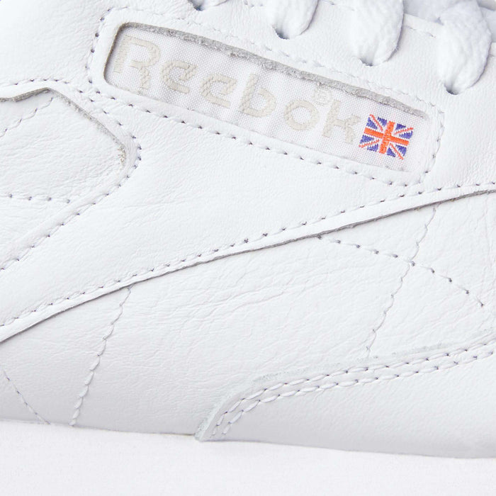 Close-up of Reebok Classic Leather Shoes in white, spotlighting the stitched Reebok logo with a Union Jack flag on the side. Features include a durable leather upper and retro-inspired stitching details.