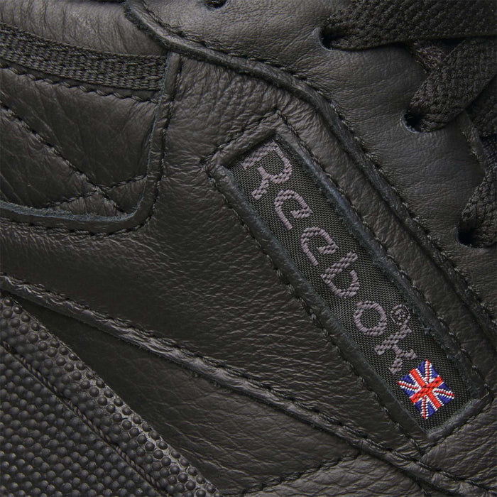Close-up of the Club C 85 Shoes by Reebok, featuring black leather, the signature Reebok logo, and a small Union Jack flag patch stitched on the side—iconic details of this classic sneaker.