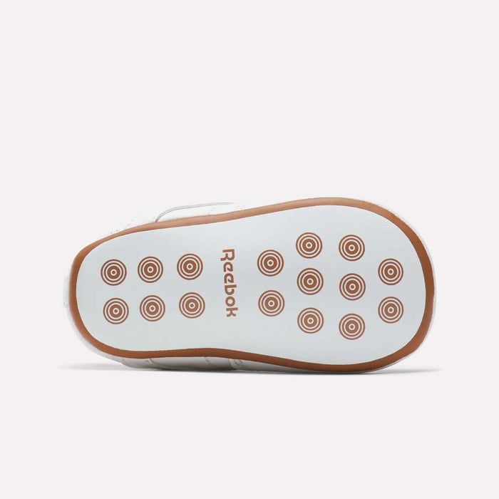 The image shows the sole of a white Kids' Club C Crib Shoe by Reebok for toddlers, featuring brown circular grip patterns and a brown Reebok logo at the center.