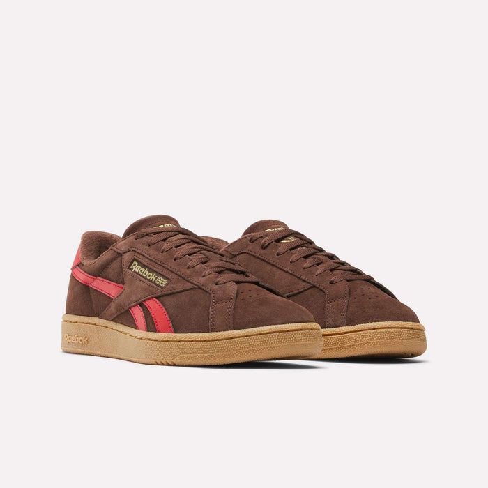 The Reebok Club C Grounds UK Shoes feature a brown suede upper, red stripes, tan soles, and gold Reebok logos on the side and tongue, displayed against a white background.