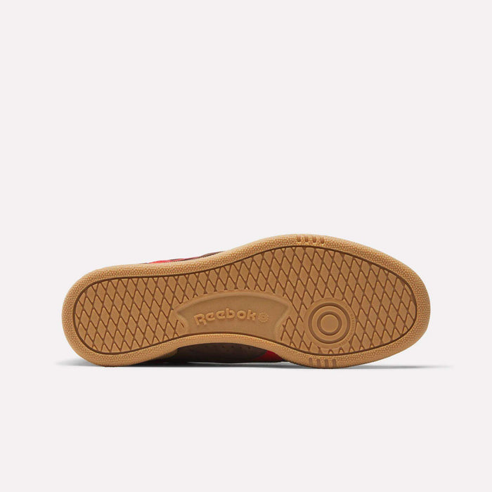 The image shows the sole of Reebok Club C Grounds UK Shoes, featuring a gum-colored rubber outsole with a circular toe pattern and herringbone grip, plus “Reebok” embossed near the arch to highlight the brand’s classic heritage.