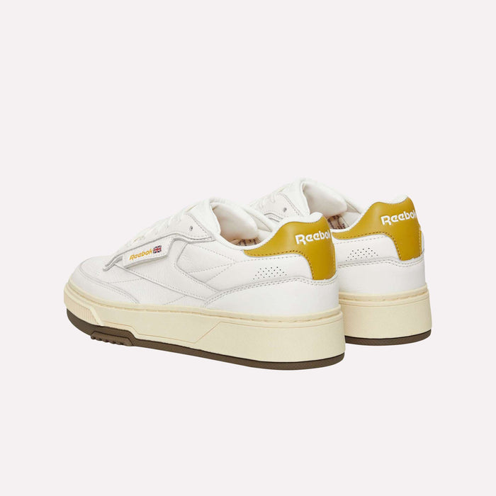 A pair of Reebok Club C LTD Shoes in white, featuring yellow accents on the heel and beige soles, are placed side by side on a white background, perfectly embodying the timeless aesthetic of a classic tennis lifestyle shoe.