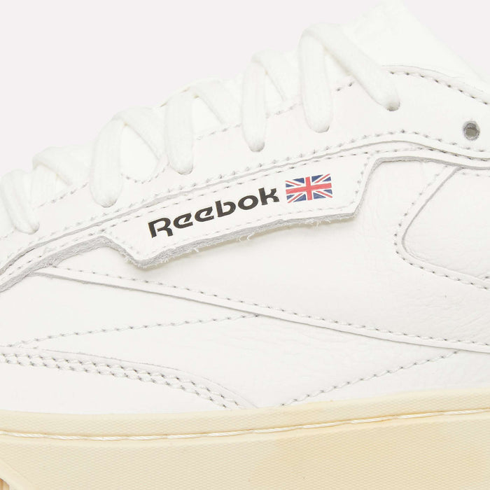 Close-up of a white Reebok Club C LTD Shoe, highlighting the Reebok logo and small British flag on the side. This iconic tennis shoe features white laces and detailed stitching atop a cream-colored sole, making it a true lifestyle classic.