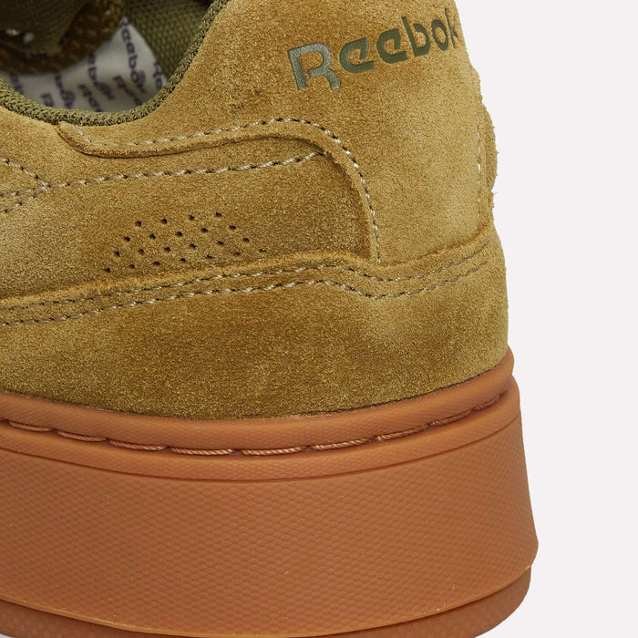 Close-up of the heel of a tan suede Reebok Club C LTD shoe featuring a light brown rubber sole. The Reebok logo appears on the upper part of this classic lifestyle tennis shoe.