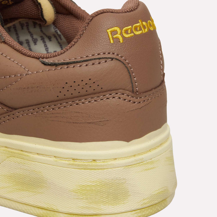 Close-up of the heel of a brown and yellow Reebok Club C LTD Shoe, featuring the embroidered Reebok logo and yellow stains on the cream-colored sole—a lifestyle classic reimagined with vintage details.