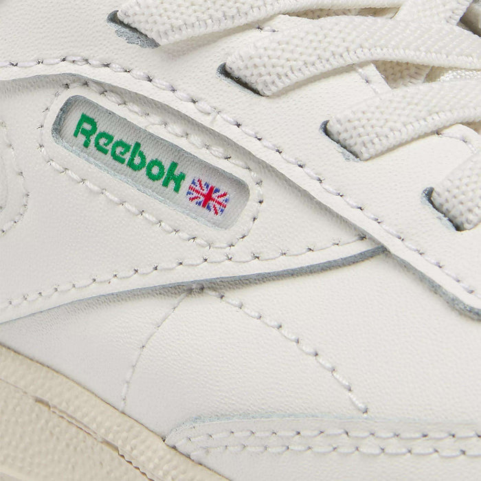 Close-up of Reebok Kids' Club C Shoes for baby & toddler in classic '80s style, featuring the green Reebok logo, small Union Jack flag on the side, visible stitching, and white laces.