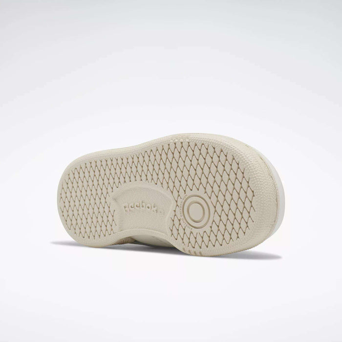 The image shows the beige sole of Reebok Kids' Club C Shoes for babies and toddlers, featuring a textured diamond pattern, circular details, and an embossed Reebok logo at the center, all set against a light background.
