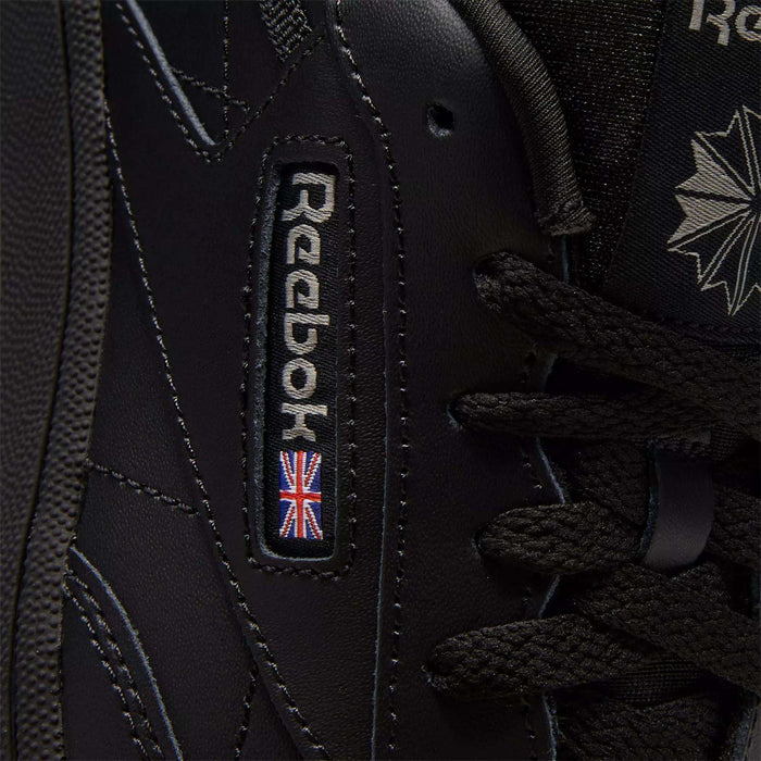 Close-up of black Reebok Kids' Club C Shoes - Grade School, showcasing classic court style with detailed stitching on soft leather uppers, plus the Reebok logo and a small UK flag patch near the laces.