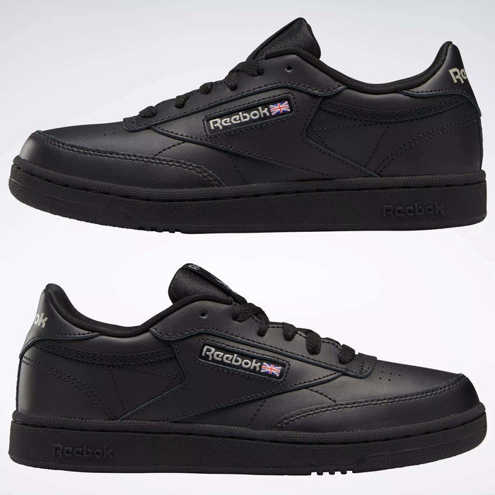 Two black Reebok Kids' Club C Shoes - Grade School are shown from the side, featuring soft leather uppers, low-top design, black soles, and the Reebok logo with a small British flag. The classic court shoe stands against a light background.